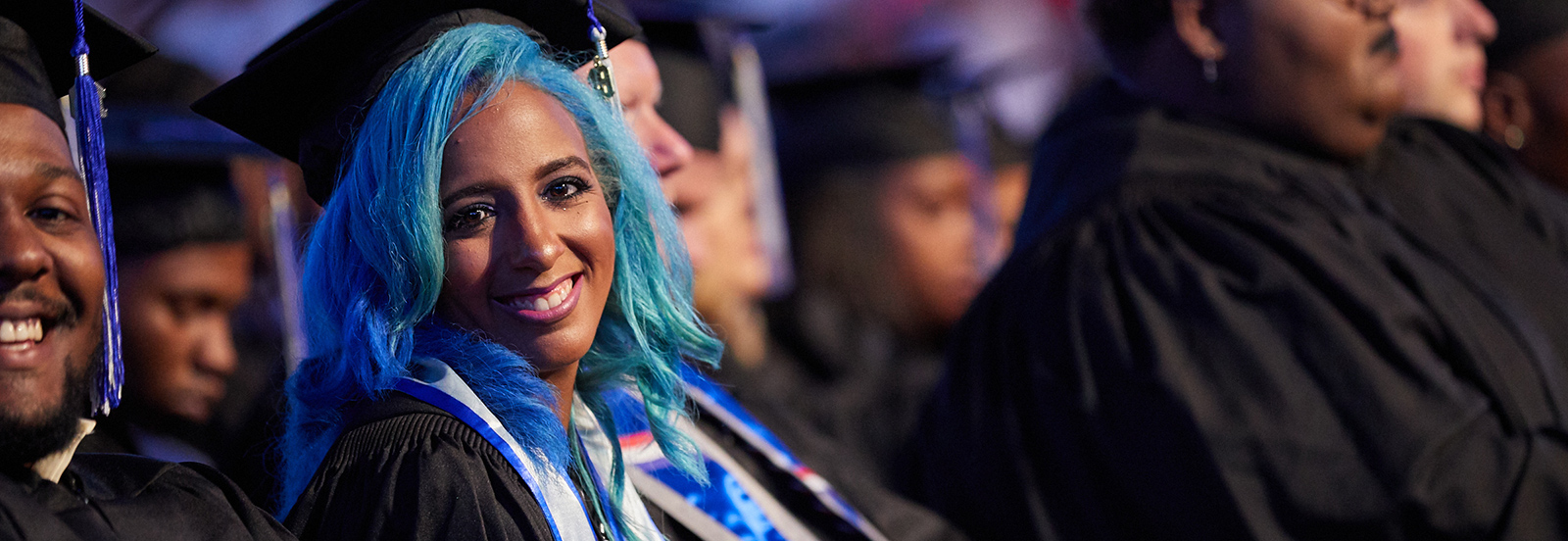 LiFE graduate with blue hair sitting in audience of graduates in commencement regalia