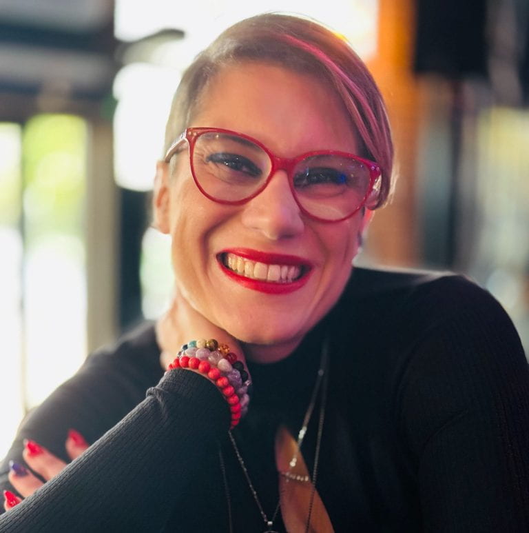 Paola Cavallari, smiles for the camera.  She is white, and wearing red glasses and lipstick. Her hair is purple. 