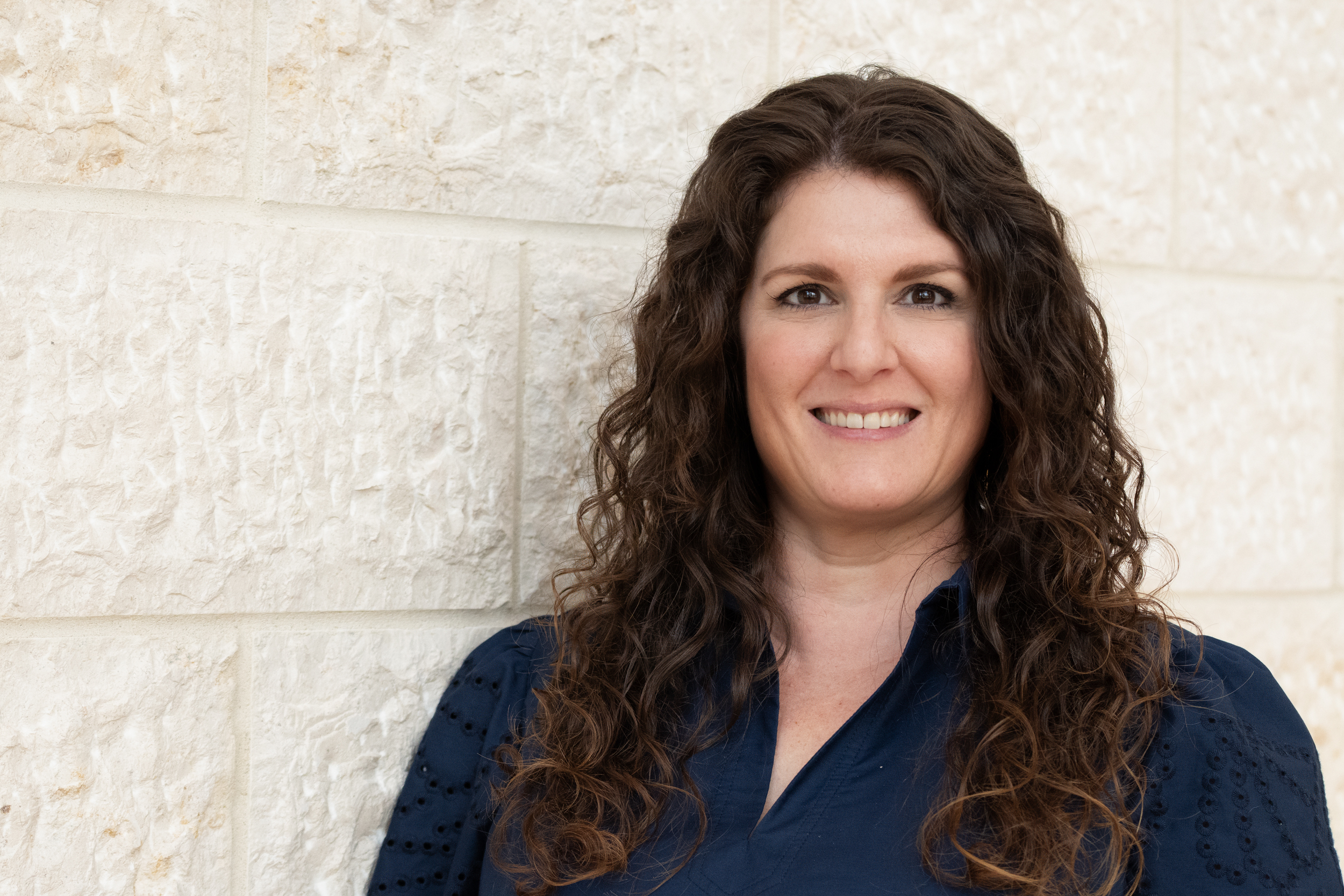 Jen Kollath, a woman with light skin and dark curly hair smiles for the camera.  She is wearing a blue blouse. 