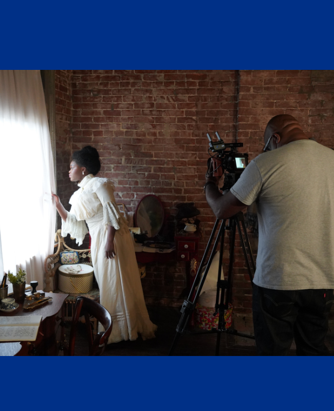 An actress dressed as Ida B Wells peers out a window curtain while a filmmaker records the scene.