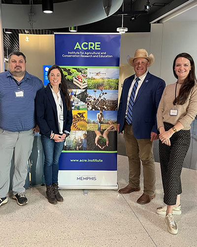 A group of people stand indoors beside a vertical banner for ACRE, the Institute of Agricultural and Conservation Research and Education. The banner features images of crops and agricultural research, along with the website “www.acre.institute” and the word “Memphis.”