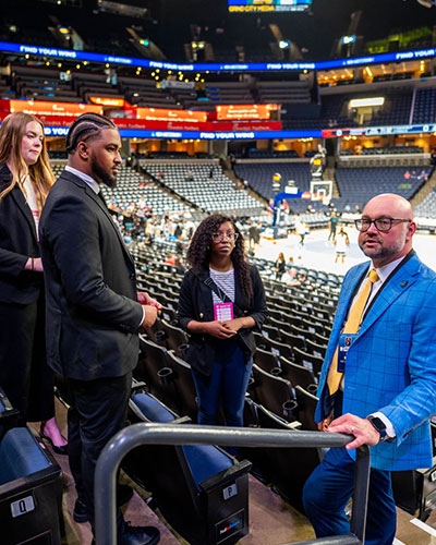 Students in business attire stand with an instructor in the seating area of FedExForum during a collegiate sports experience. Arena seats, a basketball court, and digital signage are visible in the background as the group listens and discusses.