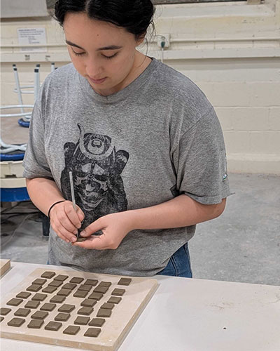 A person uses a small tool to shape square clay pieces arranged in rows on a board at a worktable. The setting appears to be a classroom or studio, with stools and equipment in the background.