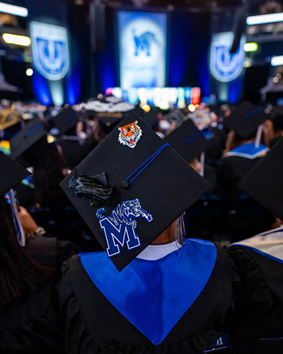 A graduate wearing a decorated cap featuring a tiger and a University of Memphis “M” sits among other graduates during a commencement ceremony. A stage with large university banners is visible in the background.