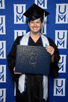April holds her diploma while smiling in her black graduation gown and cap. UofM logo wall background behind her.