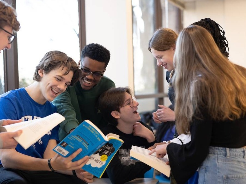 Recommended size: 800 × 600 px University High School students smiling and gathered around a classroom table with open books.