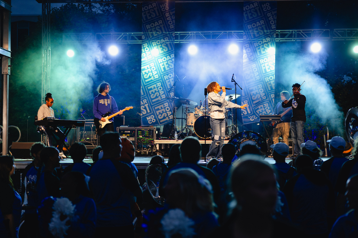 Musicians perform on an illuminated outdoor stage during University of Memphis ROAR Fest while attendees watch from the audience area.