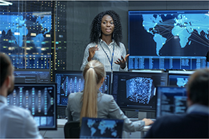 female speaking with computer monitors and screens in background