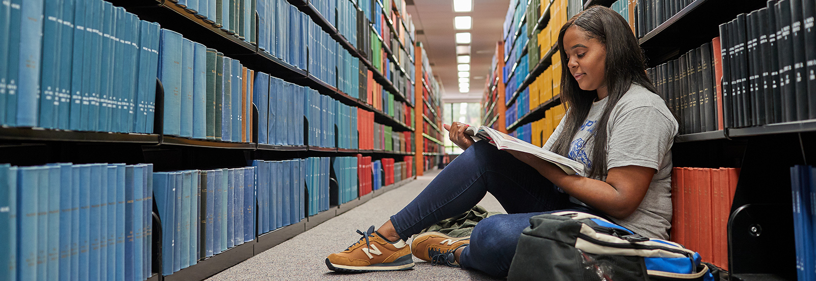 student sitting in floor of library with book shelves behind her, reading a book
