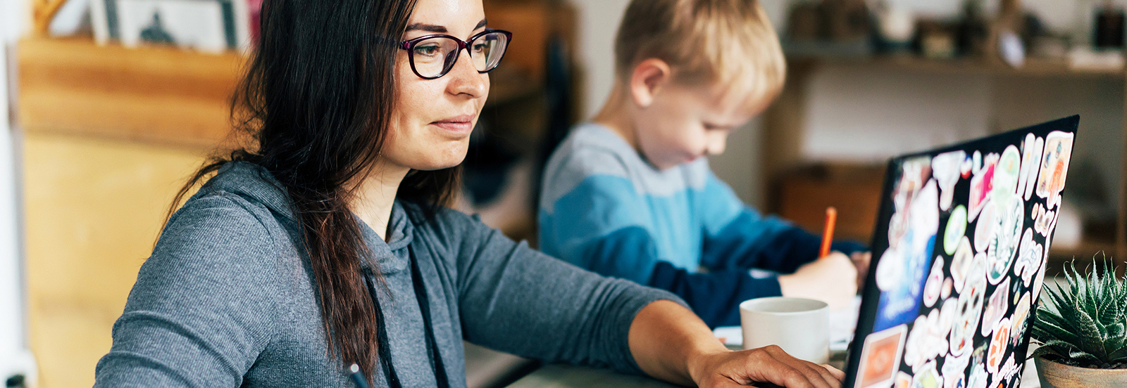 mother working on laptop with young son beside her writing with pencil on paper