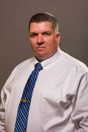 A man wearing a white dress shirt and a blue striped tie stands against a neutral background. He has short dark hair and a serious expression.