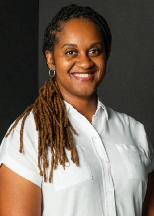 "Person standing against a dark background, wearing a white short-sleeved button-up shirt and dark green pants. They have long styled hair, blue-painted fingernails, and a smartwatch on their left wrist. The individual is smiling and looking at the camera with hands clasped in front in a relaxed pose.